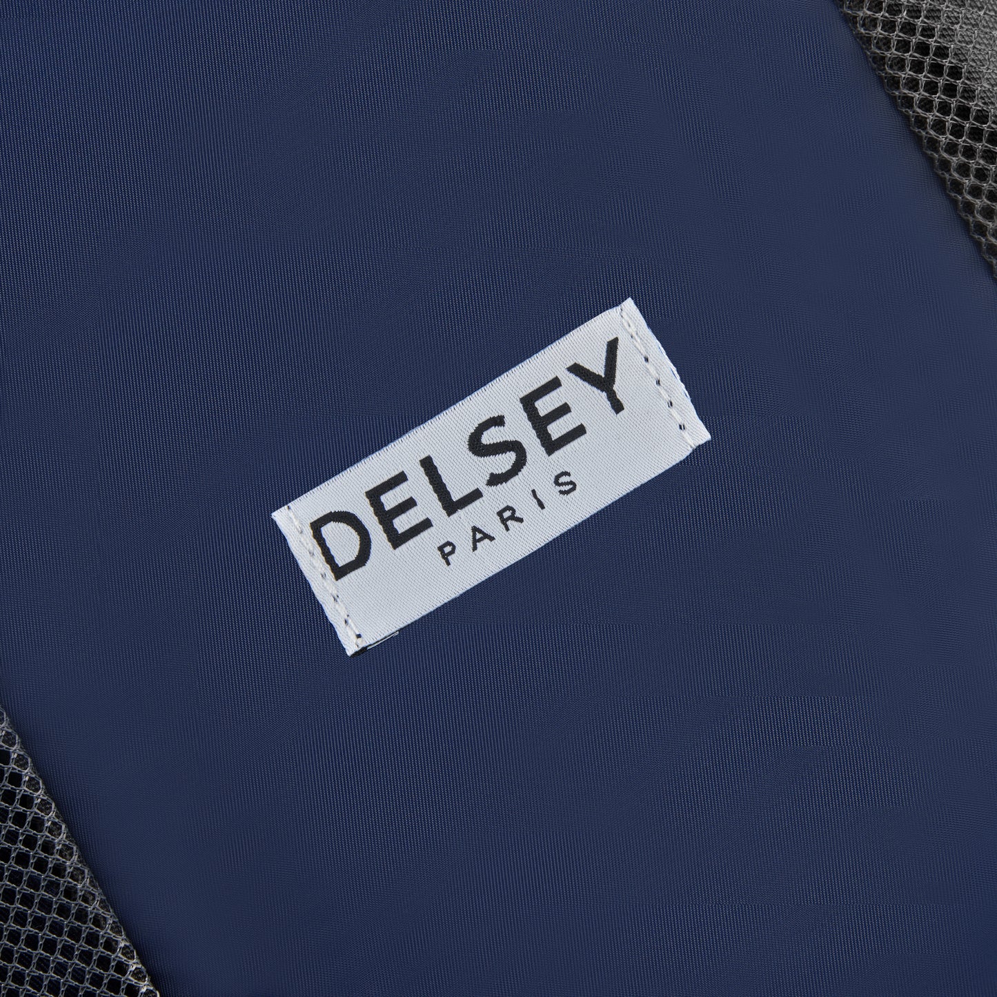 Etiqueta branca com a marca DELSEY Paris costurada em tecido azul escuro, parte de um kit organizador de viagem com acabamento em malha preta, representando acessório premium para mala de viagem.
