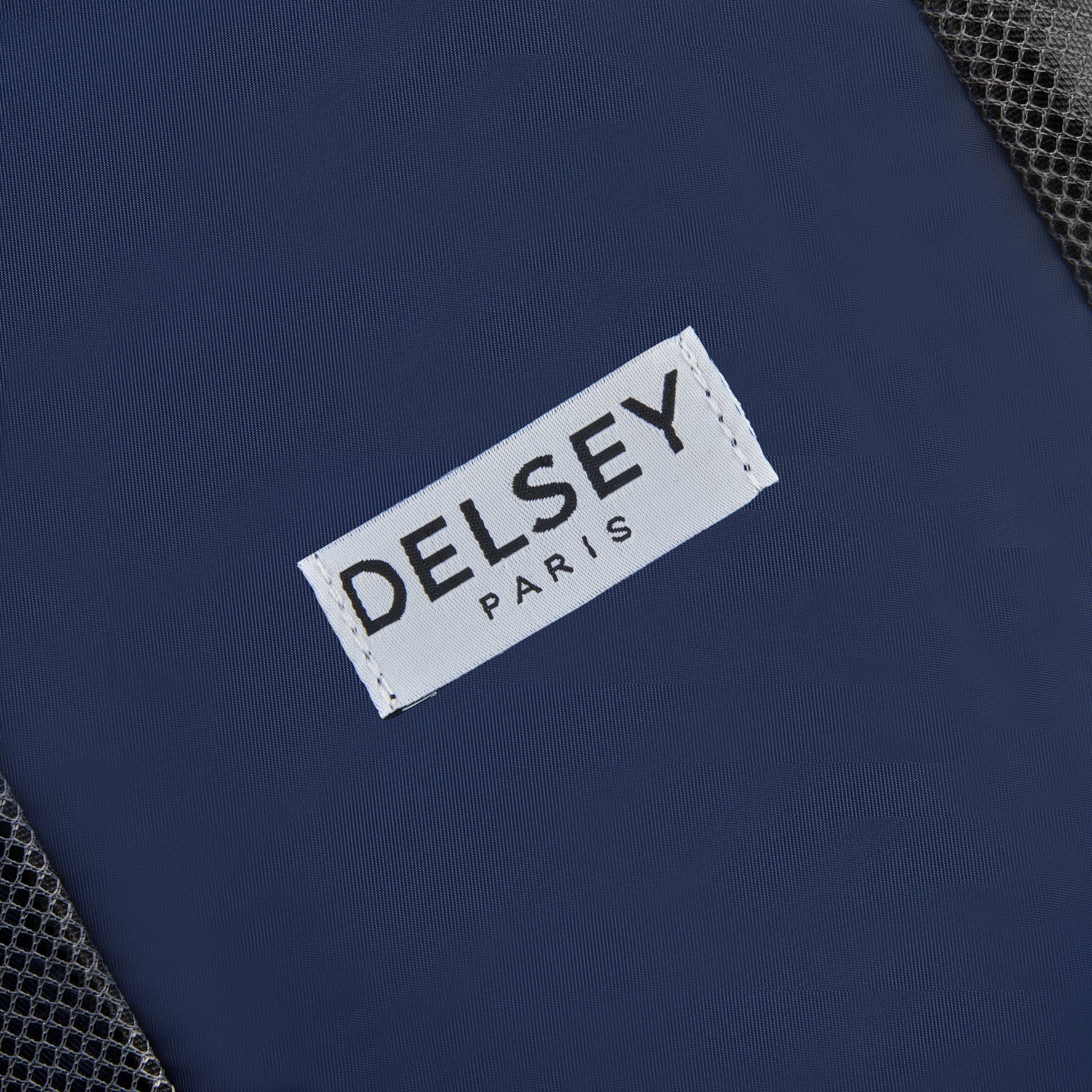 Etiqueta branca com a marca DELSEY Paris costurada em tecido azul escuro, parte de um kit organizador de viagem com acabamento em malha preta, representando acessório premium para mala de viagem.