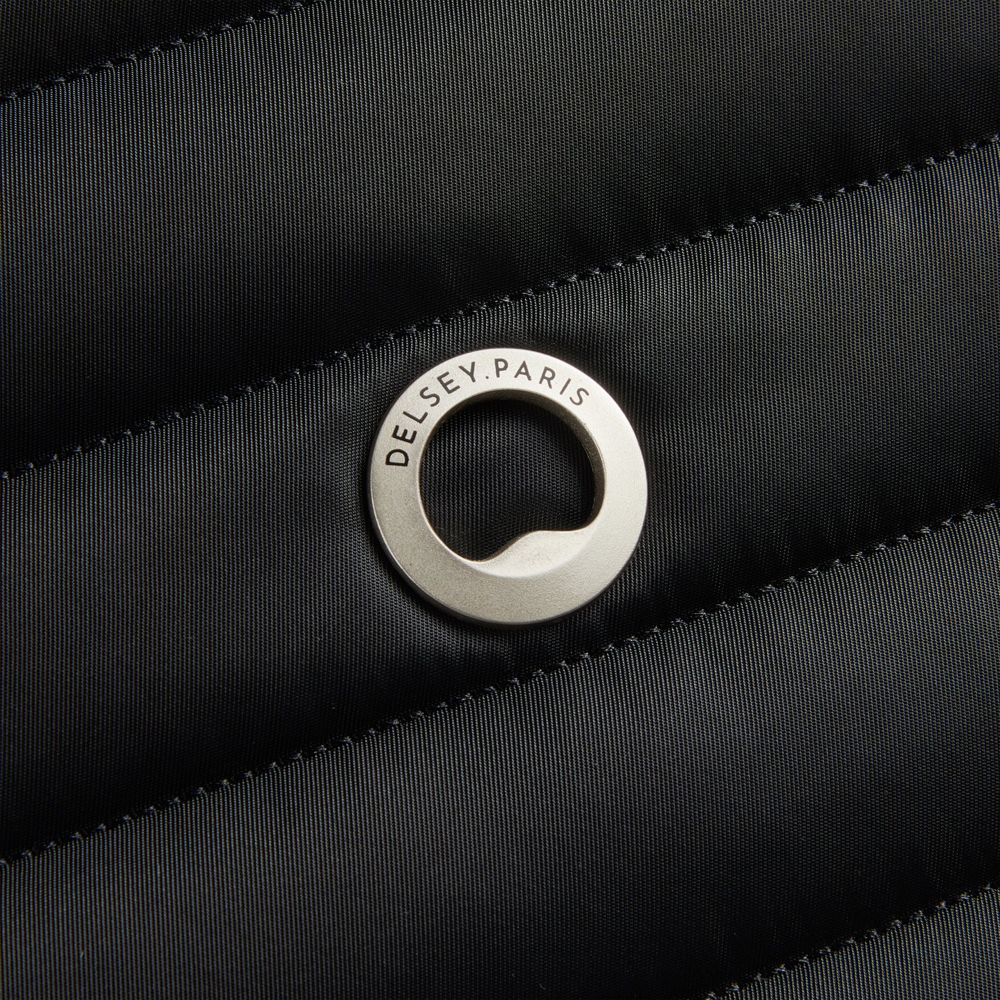 Detalhe do logo metálico da DELSEY Paris aplicado sobre tecido preto em textura acolchoada, representando a qualidade e design premium da marca.