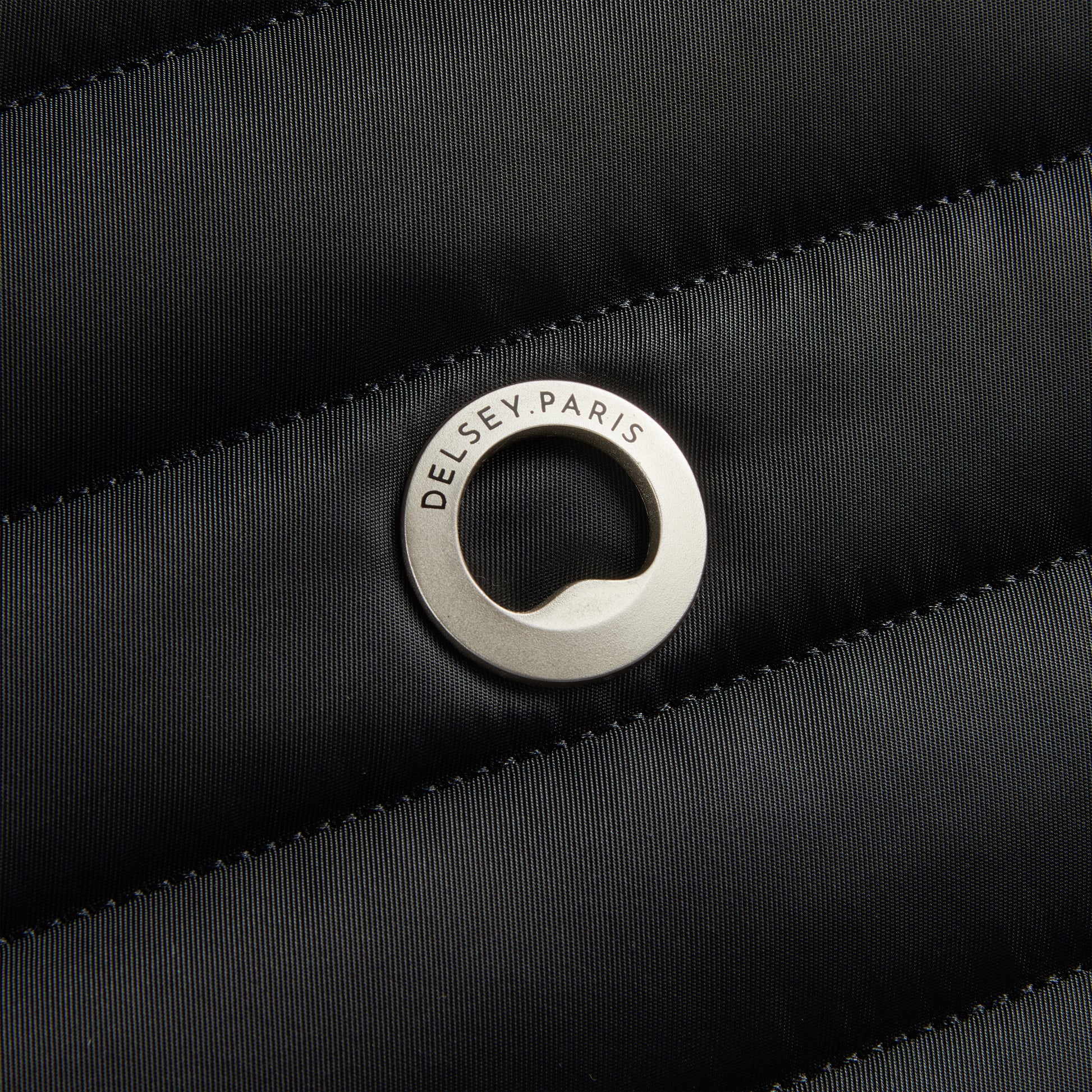 Detalhe do logo metálico da DELSEY Paris aplicado sobre tecido preto em textura acolchoada, representando a qualidade e design premium da marca.